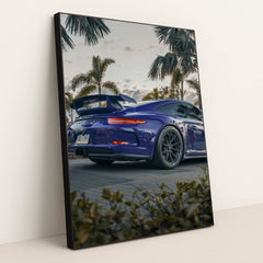 This photo shows a blue Porsche sports car with a large rear wing parked among palm trees at dusk, in a black frame.