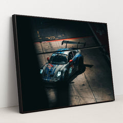 This photo shows a silver Porsche race car with blue stripes and a large rear wing in a dark garage, in a black frame.