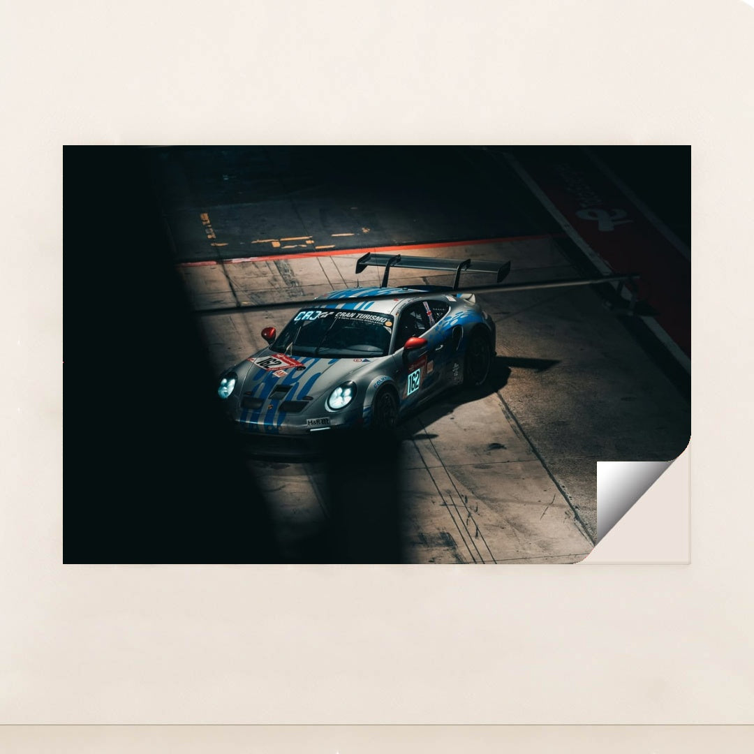 This photo shows a silver Porsche race car with blue stripes and a large rear wing in a dark garage, unframed.