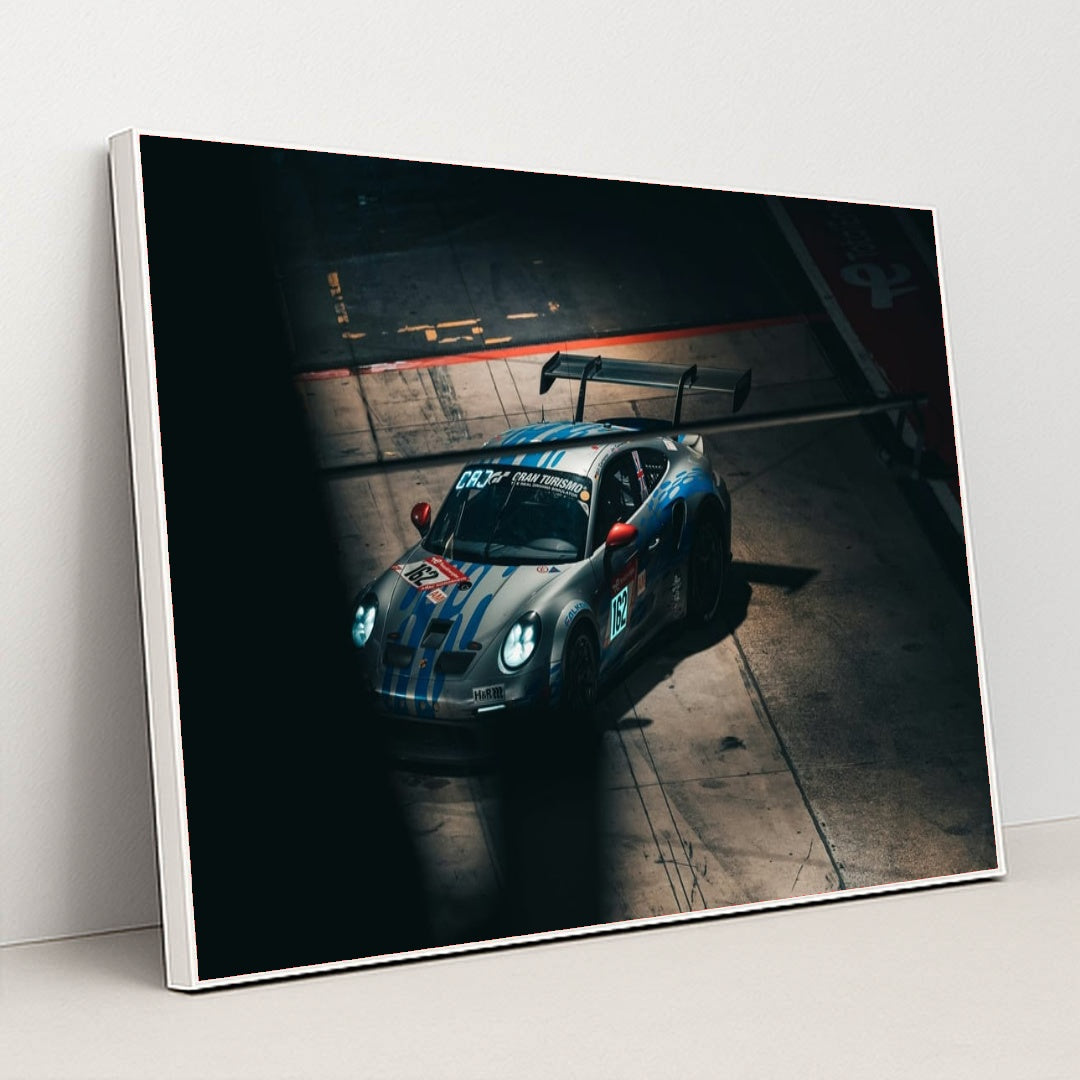This photo shows a silver Porsche race car with blue stripes and a large rear wing in a dark garage, in a white frame.