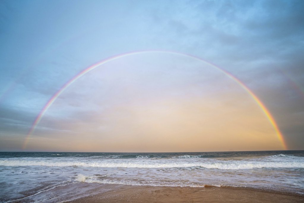 Ben Mack - A stunning rainbow arches over a serene beachfront at sunset, creating a tranquil seaside atmosphere.