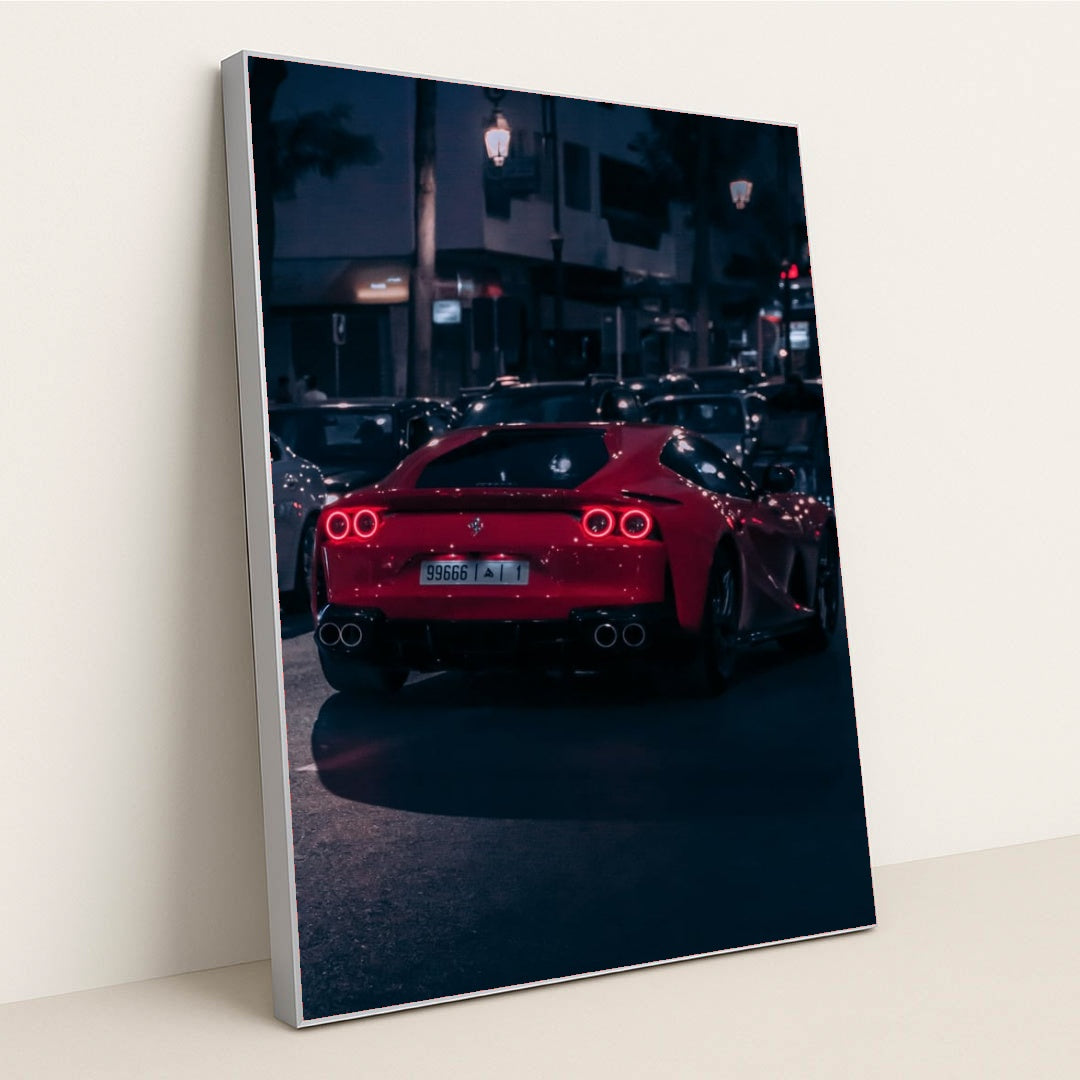 This photo shows a red Ferrari sports car driving at night through city traffic under streetlights, in a silver frame.