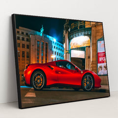 This photo shows a red Ferrari sports car parked in front of an ornate city building at night, under bright streetlights, in a black frame.
