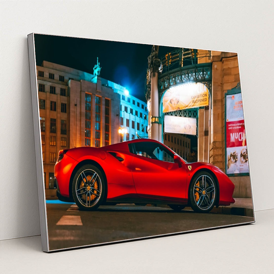 This photo shows a red Ferrari sports car parked in front of an ornate city building at night, under bright streetlights, in a silver frame.