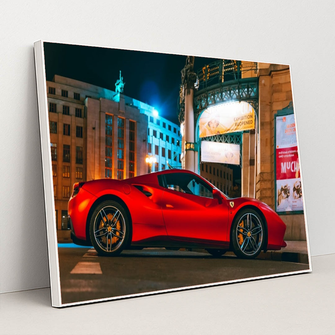 This photo shows a red Ferrari sports car parked in front of an ornate city building at night, under bright streetlights, in a white frame.