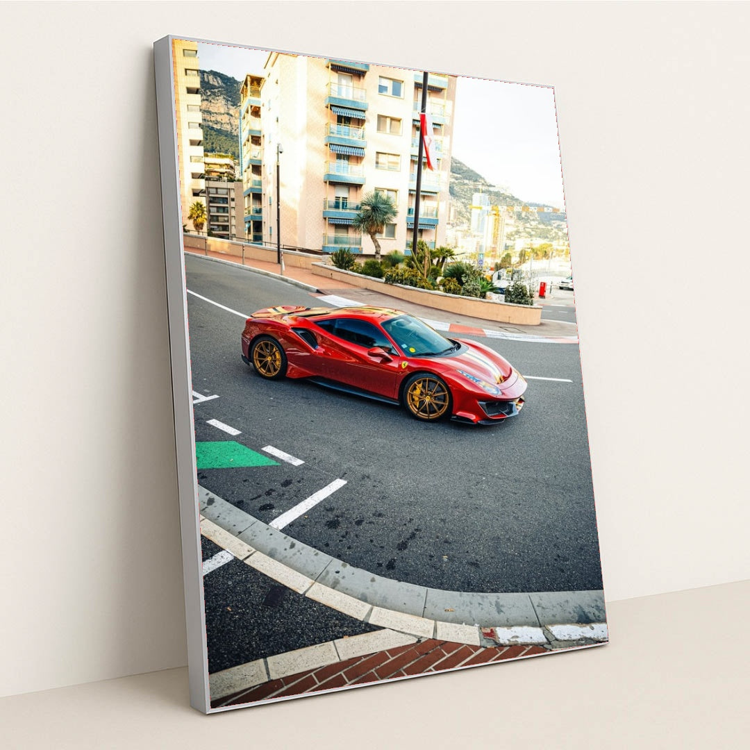 This photo shows a red Ferrari 488 Pista with gold wheels driving through a city street with Mediterranean buildings, in a silver frame.