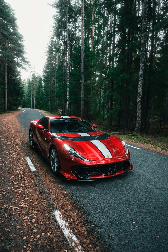 Ferrari 812 Superfast on the Forest Path, a metallic red car on a winding road surrounded by tall trees and fallen leaves.