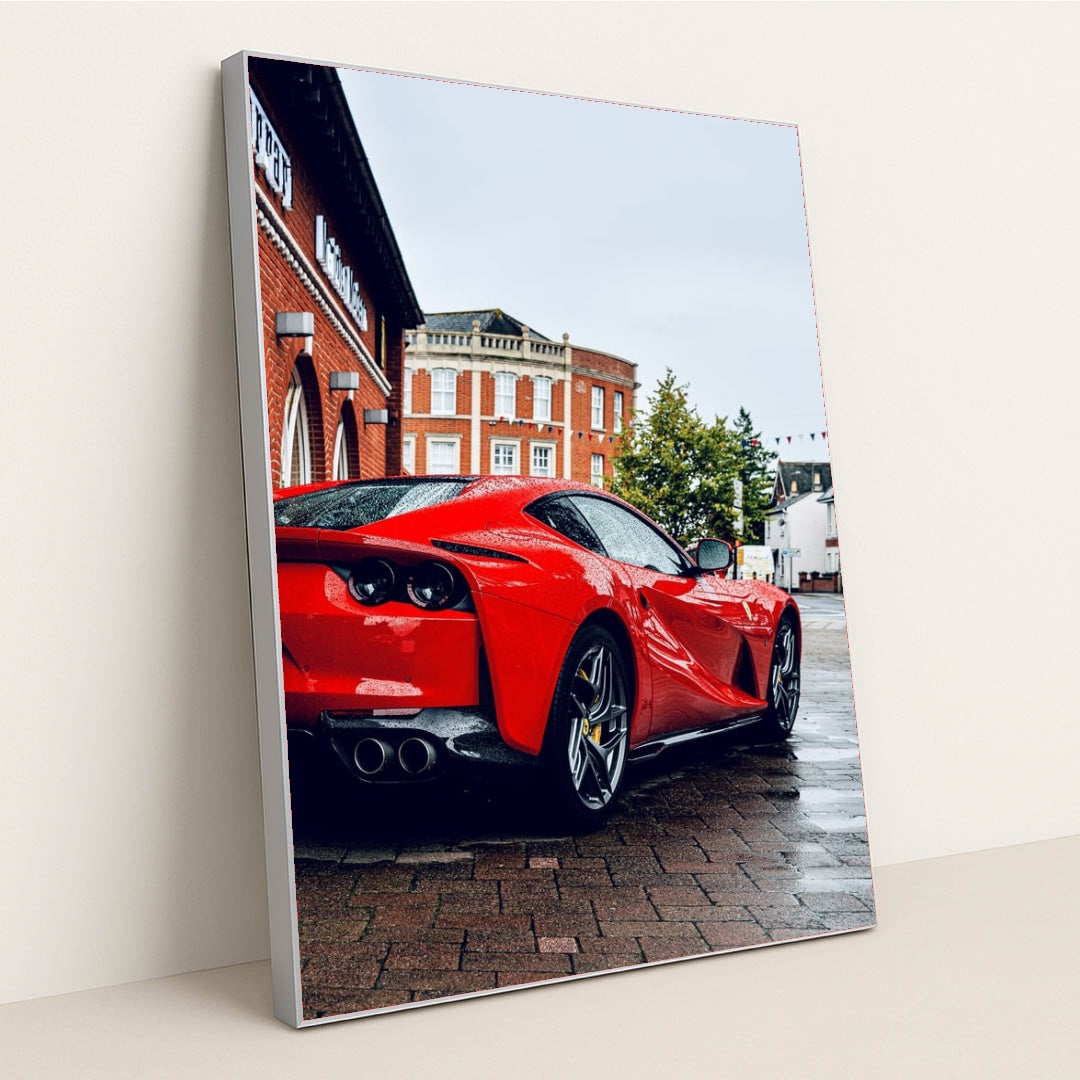 This photo shows the rear three-quarter view of a red Ferrari 812 Superfast parked on a wet city street, in a silver frame.