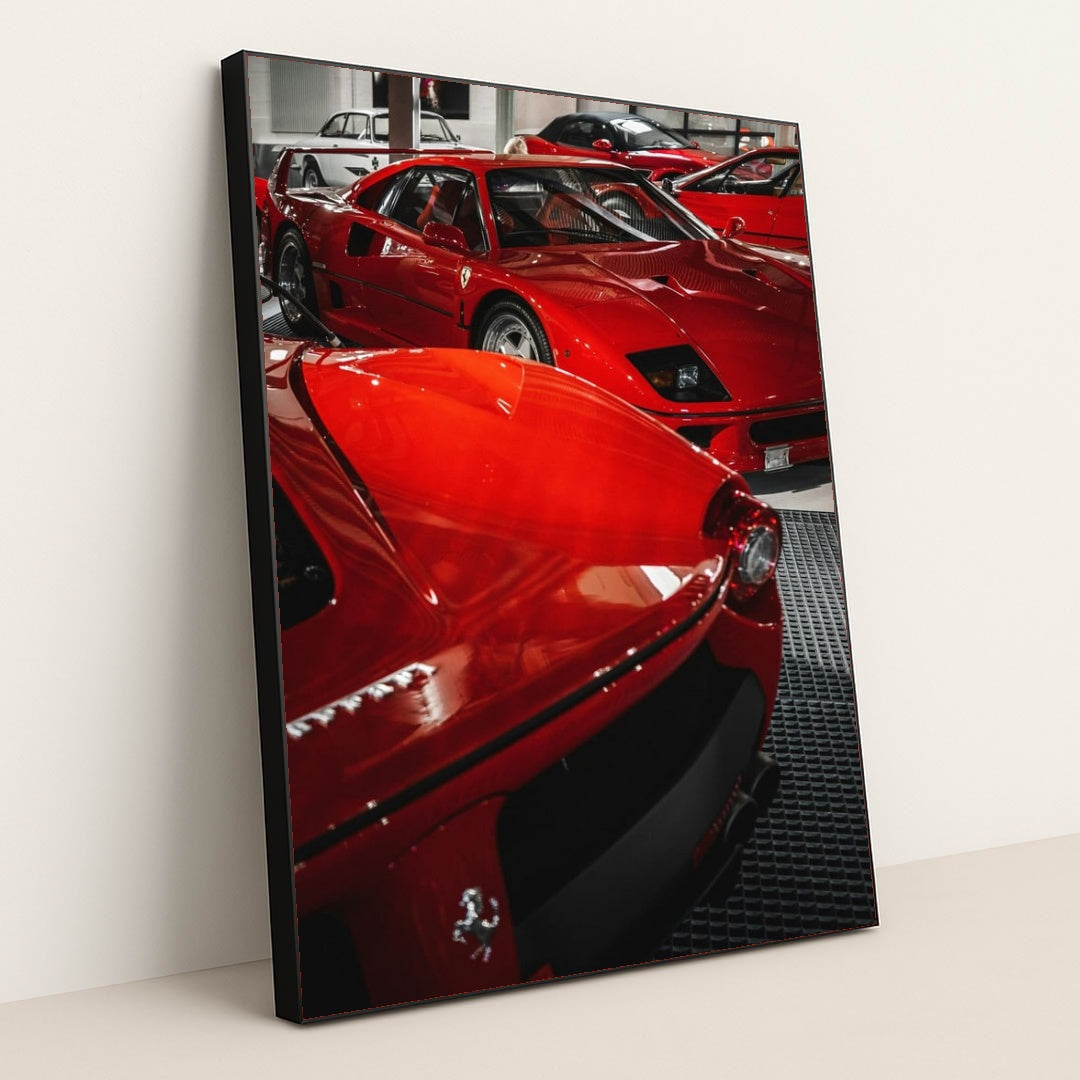 This photo shows a group of classic red Ferrari supercars displayed indoors on a black grid floor, in a black frame.