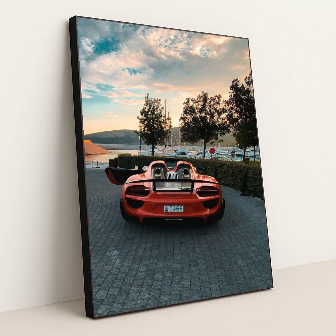 This photo shows a red Porsche 918 Spyder with open doors parked on a cobbled marina at sunset, in a black frame.