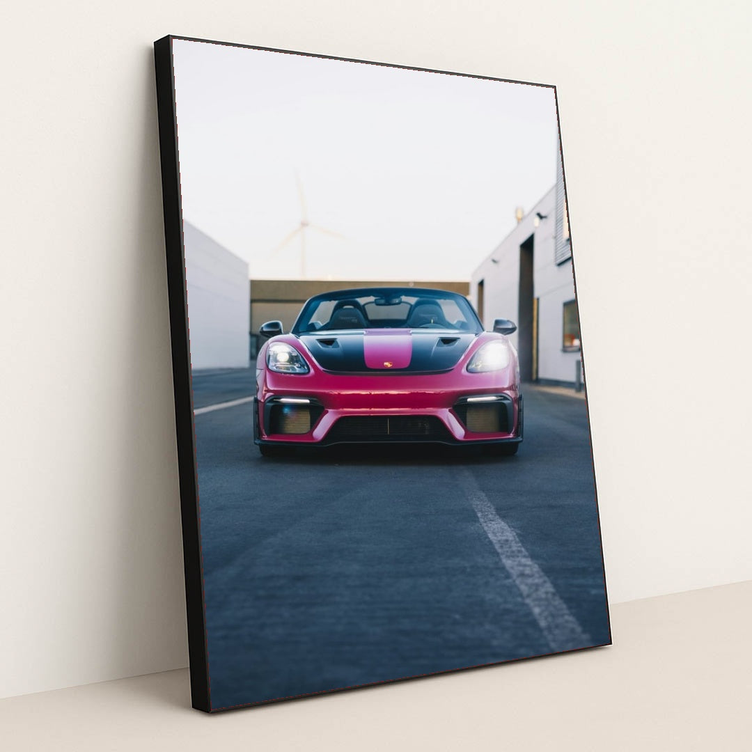 This photo shows a pink and black Porsche sports car viewed from the front, parked on an urban street, in a black frame.