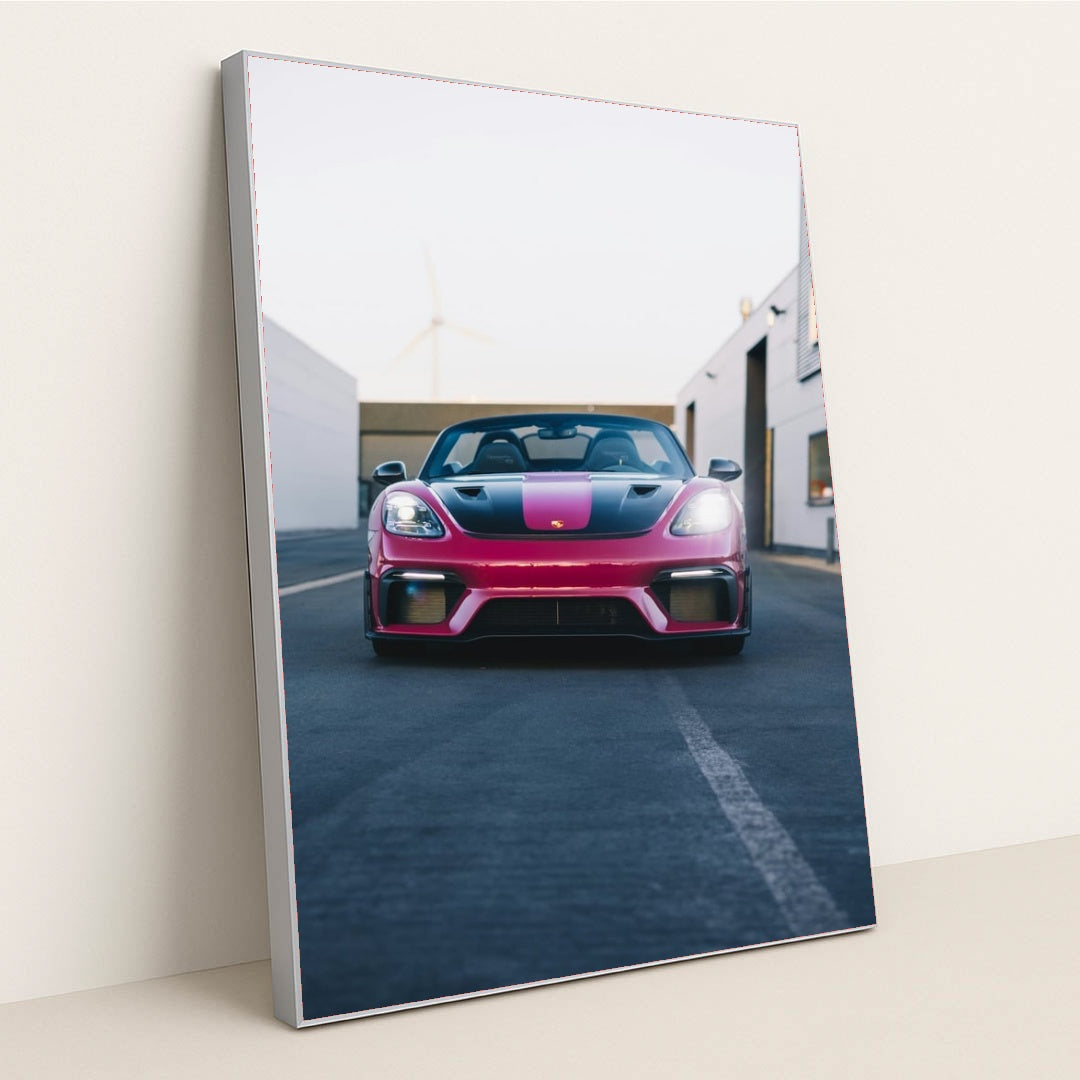 This photo shows a pink and black Porsche sports car viewed from the front, parked on an urban street, in a silver frame.