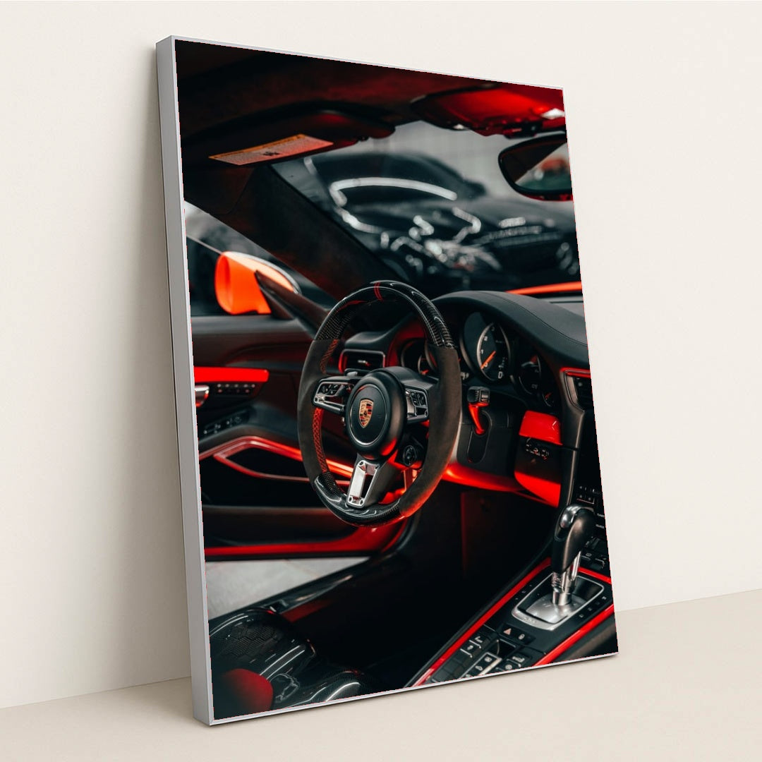 This photo shows the interior of a Porsche with a black and red steering wheel, paddle shifters, and orange accents, in a silver frame.