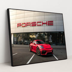 This photo shows a red Porsche sports car parked in front of a modern dealership building with a large Porsche sign, in a black frame.