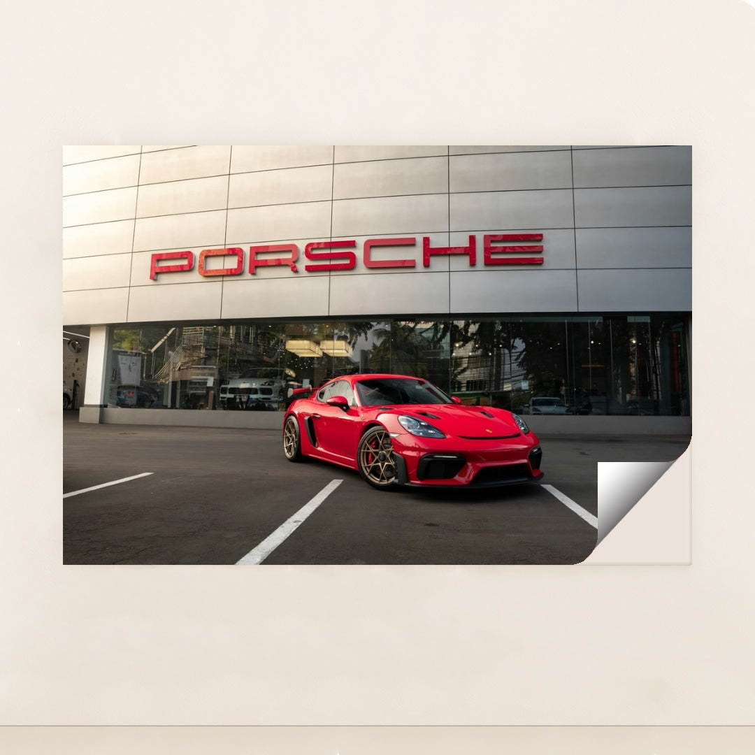 This photo shows a red Porsche sports car parked in front of a modern dealership building with a large Porsche sign, unframed style.