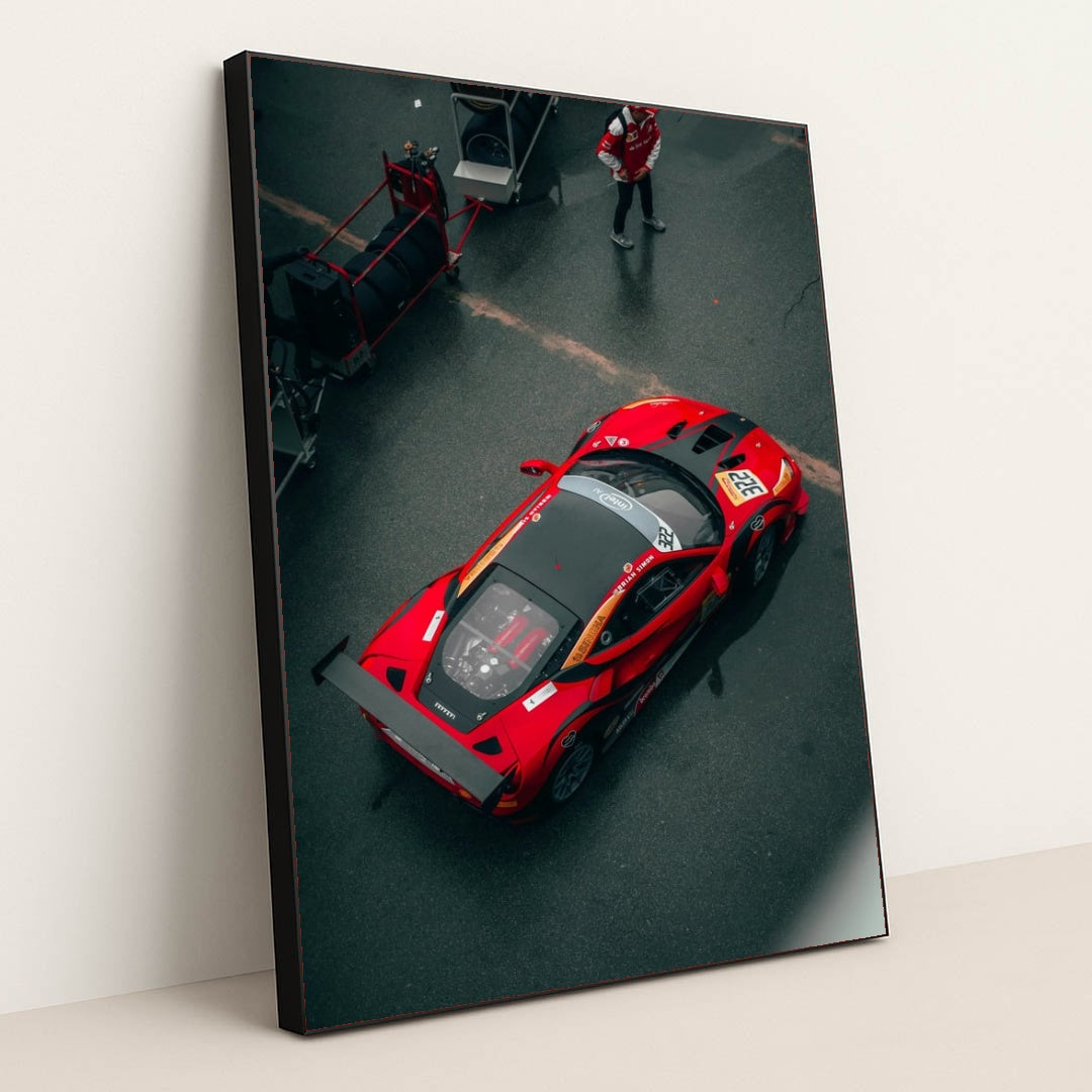 This photo shows an overhead view of a red Ferrari race car with a large rear wing parked in the pit lane, in a black frame.