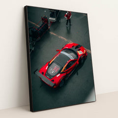This photo shows an overhead view of a red Ferrari race car with a large rear wing parked in the pit lane, in a black frame.