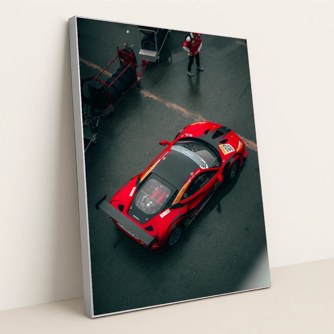 This photo shows an overhead view of a red Ferrari race car with a large rear wing parked in the pit lane, in a silver frame.