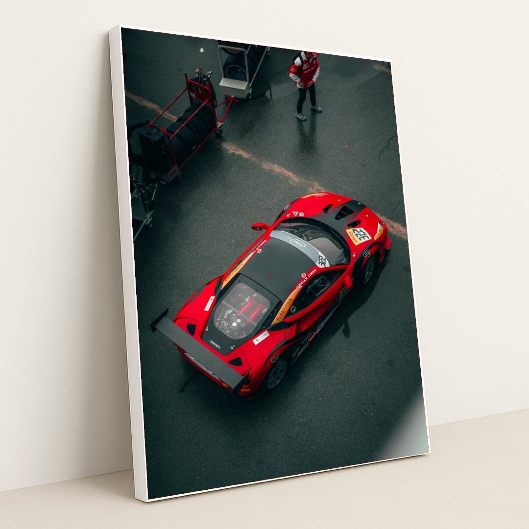 This photo shows an overhead view of a red Ferrari race car with a large rear wing parked in the pit lane, in a white frame.