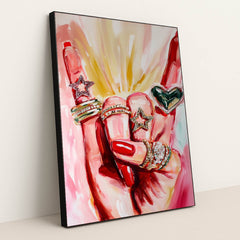 This artwork shows a hand with red nails wearing sparkly rings shaped like stars and a heart, set against pink painted brushstrokes, in a black frame.