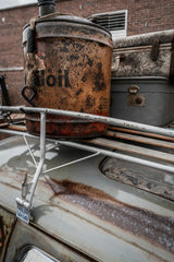 Connor McManus - Close-up of a rusty oil can and luggage on a vintage car roof with urban background.