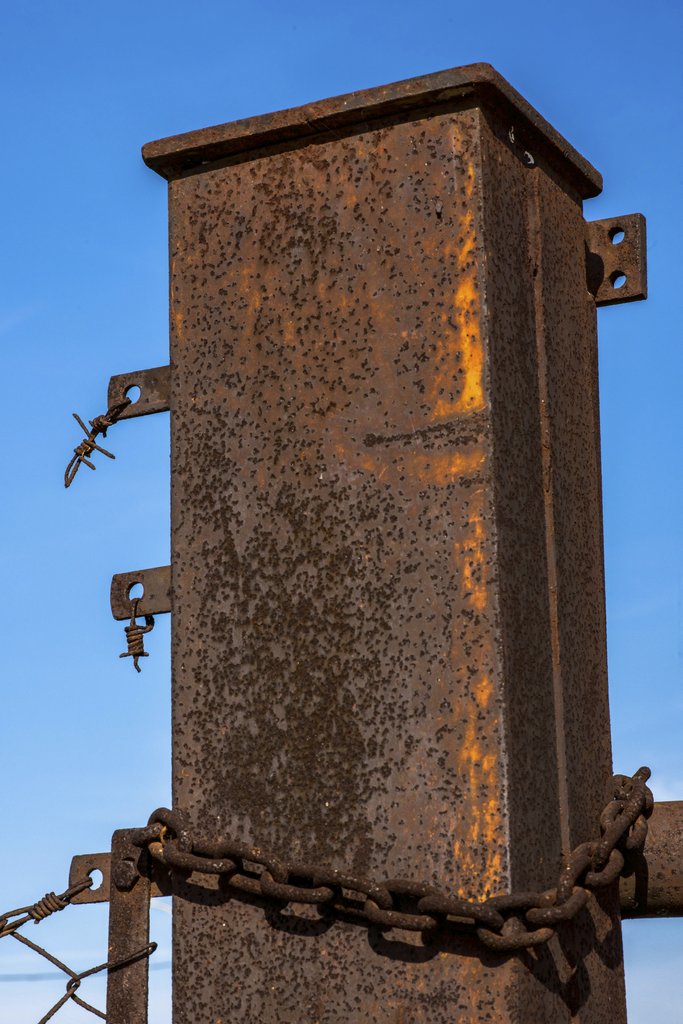 Victor  Moragriega - Close-up of a rusted metal post with chains and barbed wire against a clear blue sky.