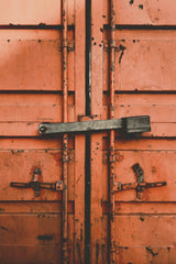 David Underland - Detailed view of weathered, orange metal container doors secured with a black bar.