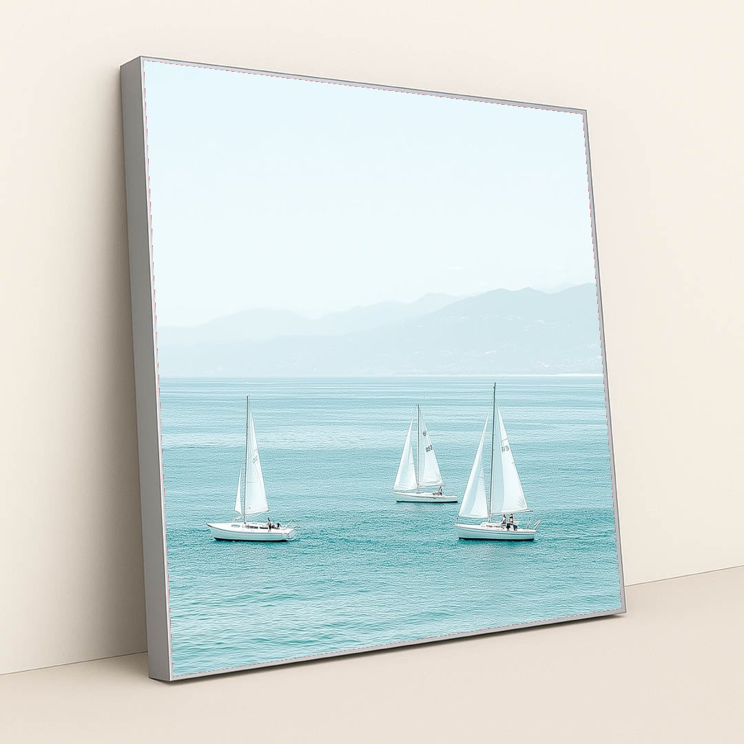 This artwork shows three white sailboats on calm turquoise water with mountains in the distance, in a silver frame.