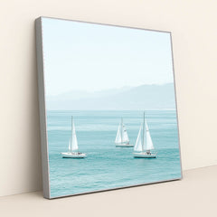 This artwork shows three white sailboats on calm turquoise water with mountains in the distance, in a silver frame.