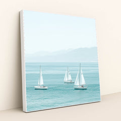 This artwork shows three white sailboats on calm turquoise water with mountains in the distance, in a white frame.