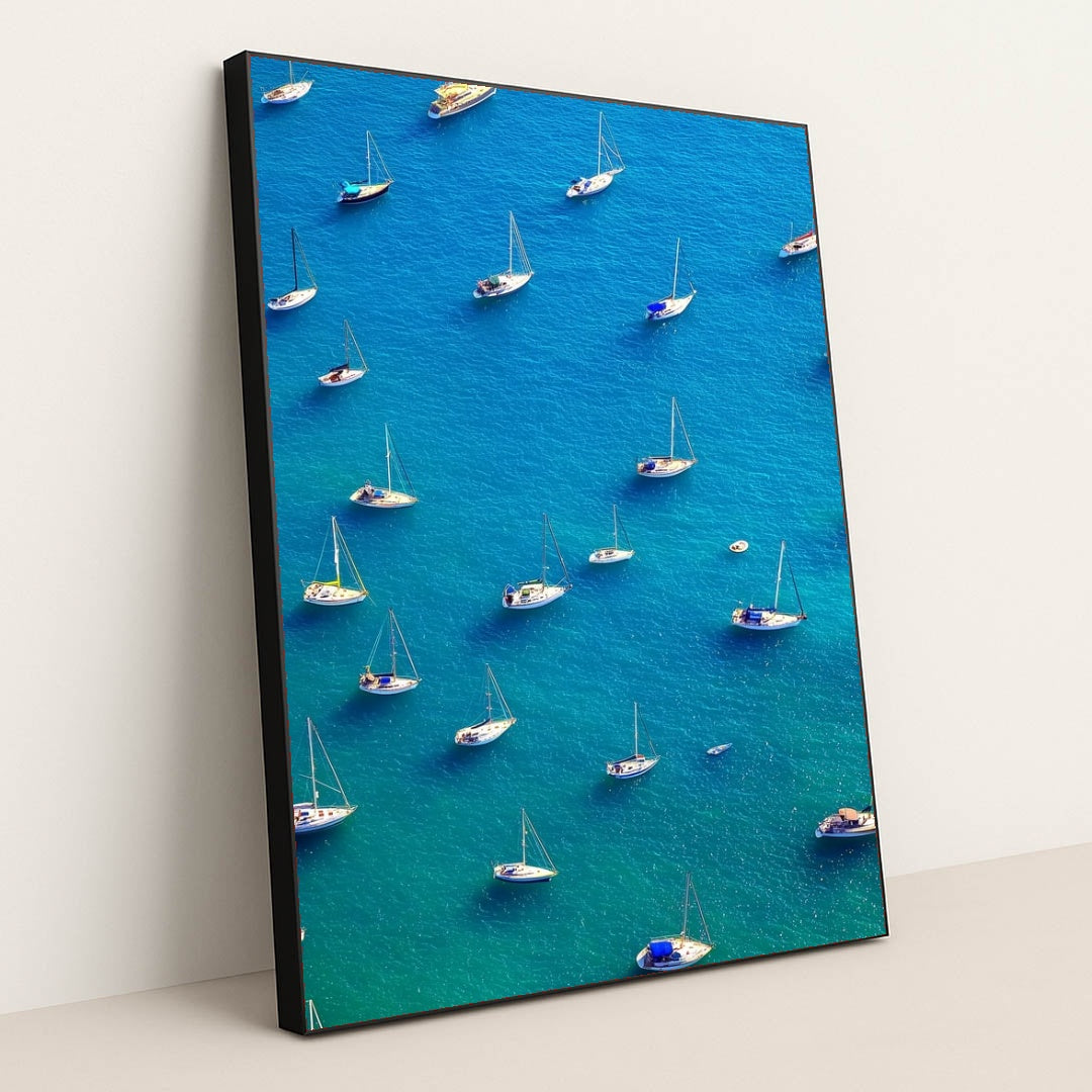 This artwork features an aerial view of moored sailboats on bright blue water, in a black frame.