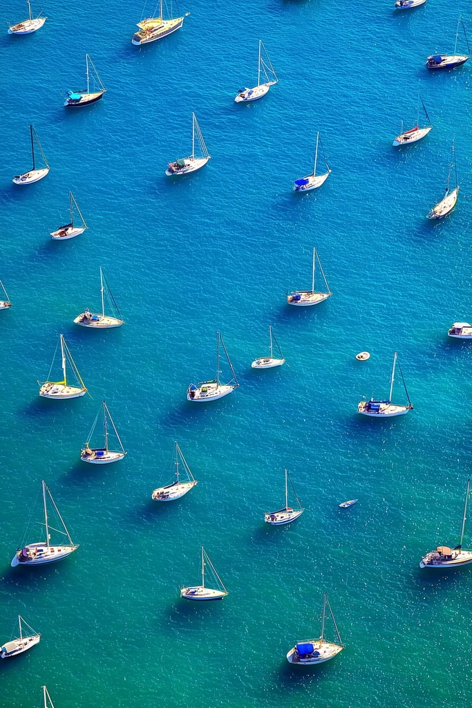 This artwork features an aerial view of moored sailboats on bright blue water.