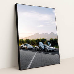 This photo shows a white Porsche sports car and a silver supercar with open butterfly doors parked by the mountains at sunrise, in a black frame.