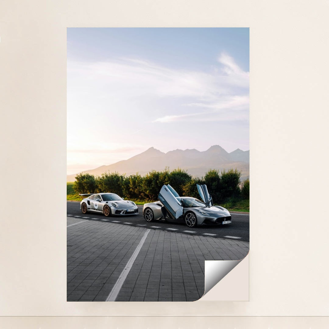 This photo shows a white Porsche sports car and a silver supercar with open butterfly doors parked by the mountains at sunrise, unframed.