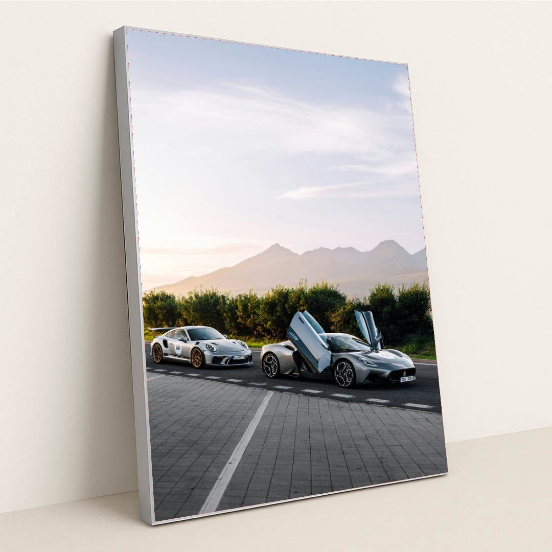 This photo shows a white Porsche sports car and a silver supercar with open butterfly doors parked by the mountains at sunrise, in a silver frame.