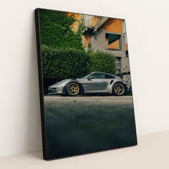 This photo shows a silver Porsche 911 GT3 RS with gold wheels parked beside a hedge and brick building, in a black frame.