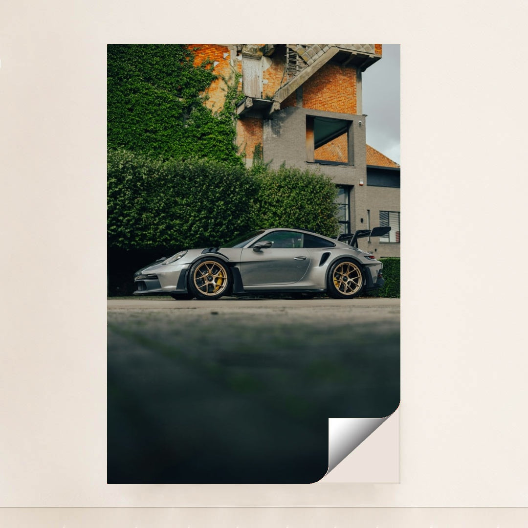 This photo shows a silver Porsche 911 GT3 RS with gold wheels parked beside a hedge and brick building, unframed style.