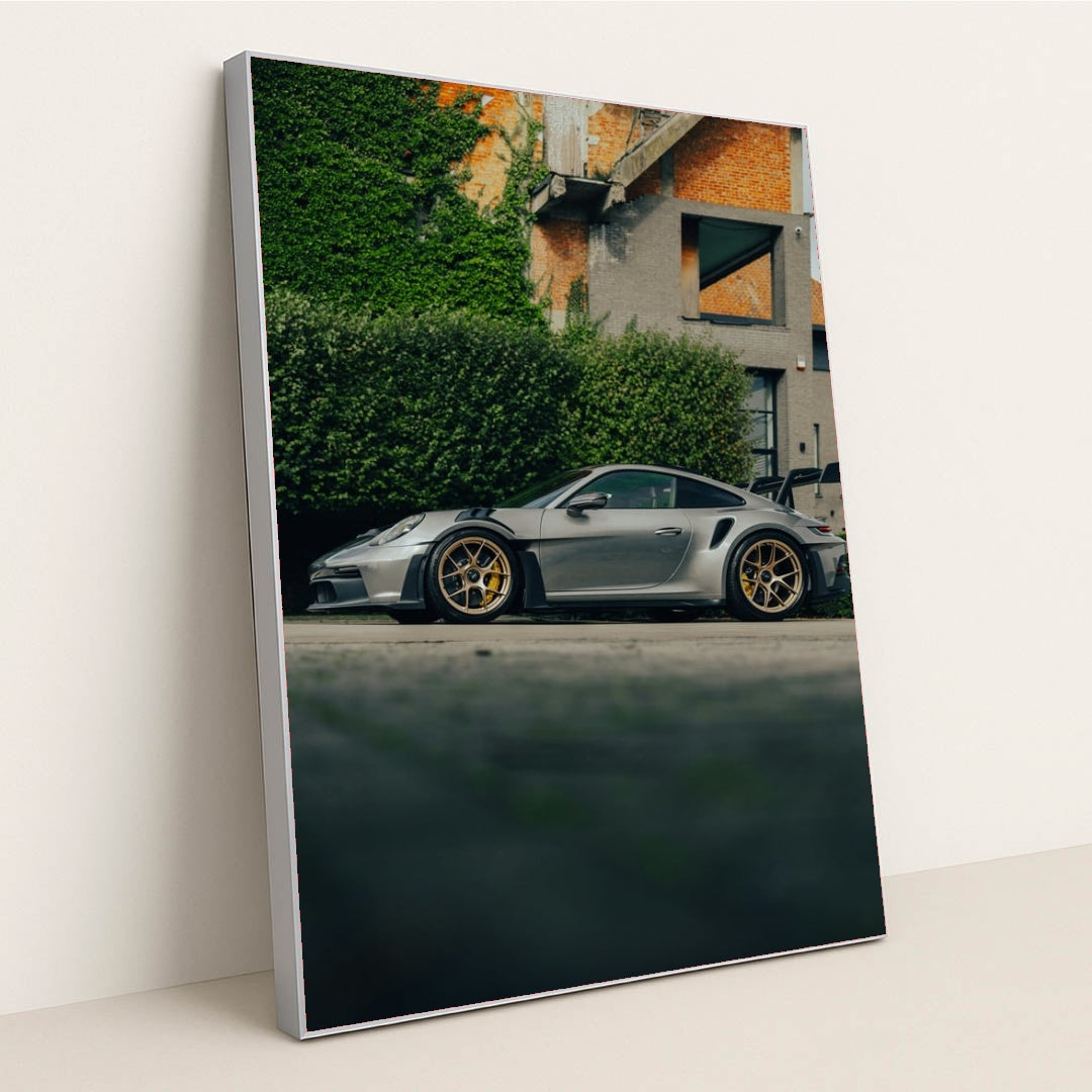 This photo shows a silver Porsche 911 GT3 RS with gold wheels parked beside a hedge and brick building, in a silver frame.