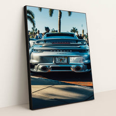 This photo shows the rear of a silver Porsche 911 Turbo S parked outdoors with palm trees and people in the background, in a black frame.