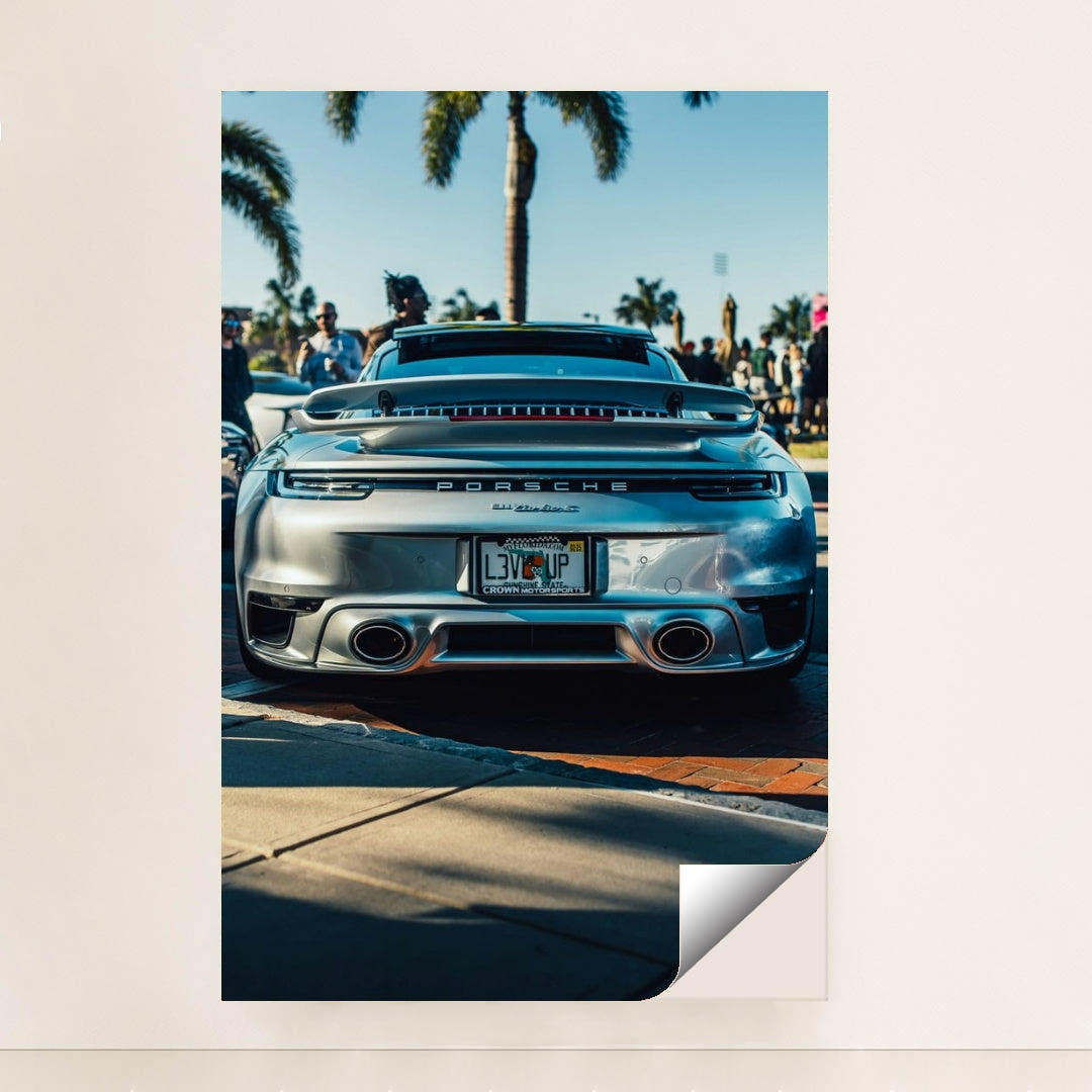 This photo shows the rear of a silver Porsche 911 Turbo S parked outdoors with palm trees and people in the background, unframed.