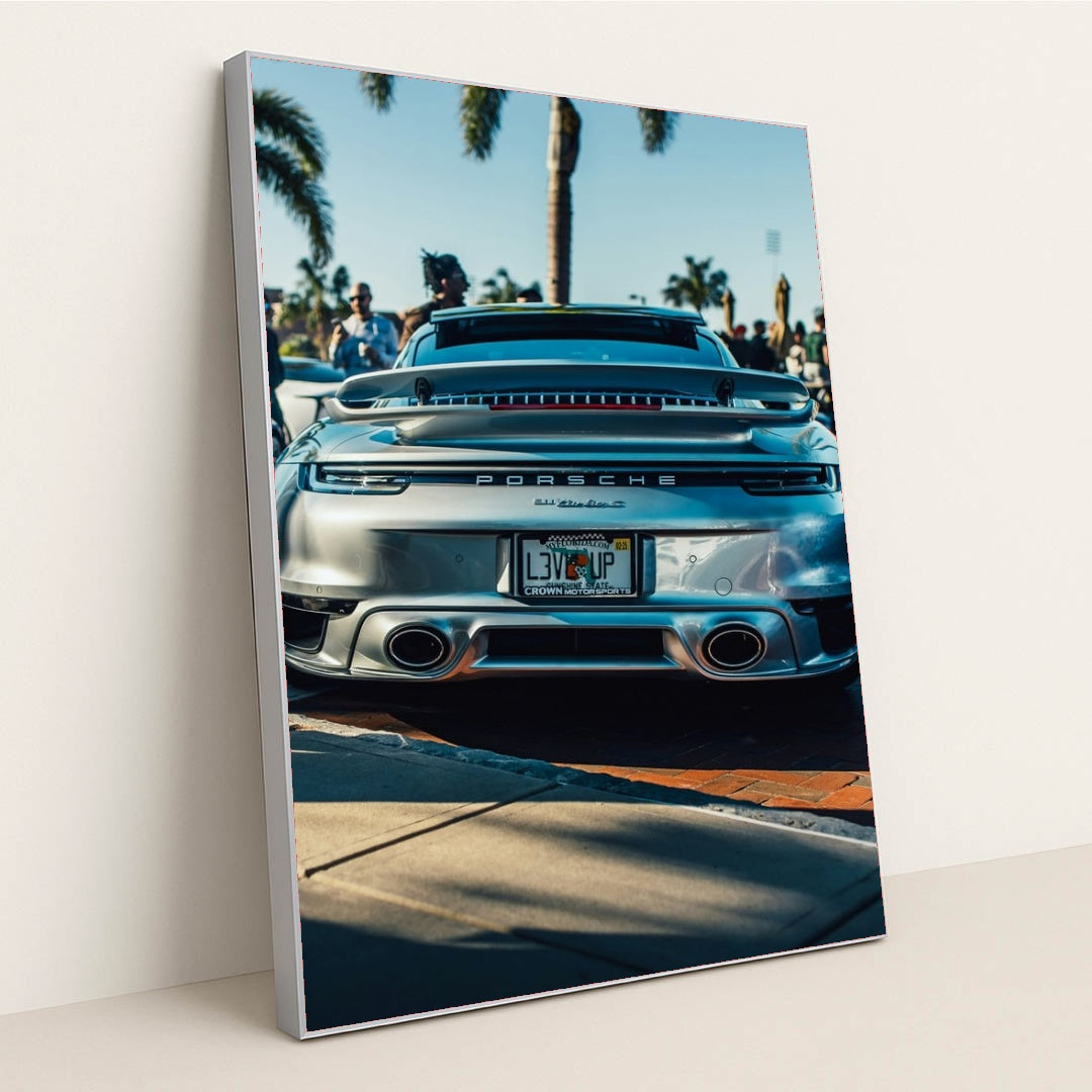 This photo shows the rear of a silver Porsche 911 Turbo S parked outdoors with palm trees and people in the background, in a silver frame.