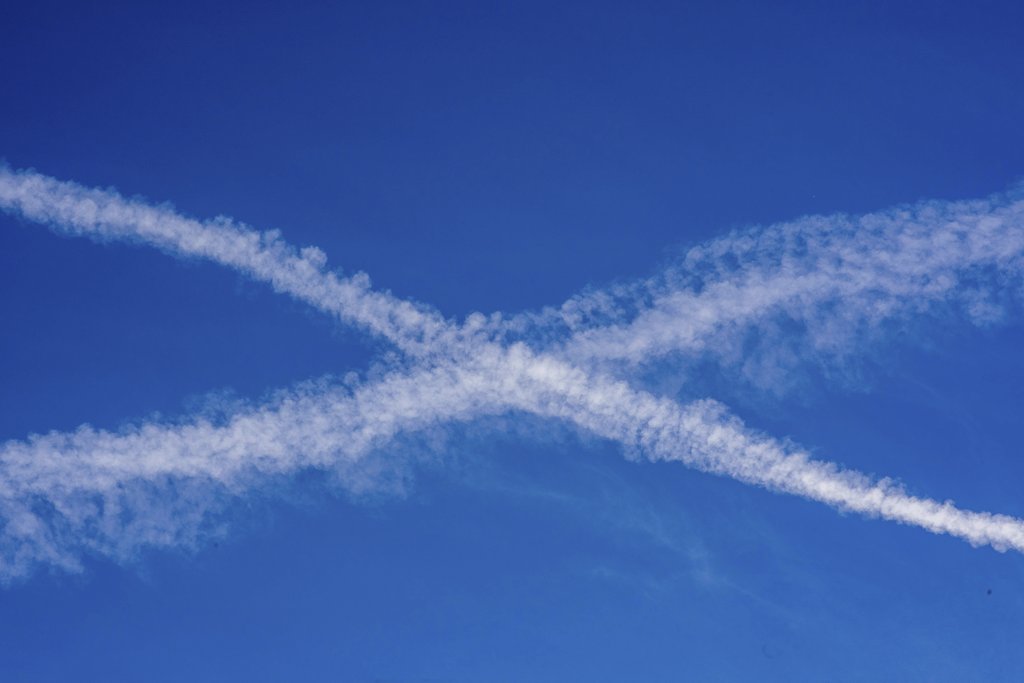 Victor  Moragriega - Airplane contrails forming a cross pattern in a vibrant blue sky, showing dynamic movement.