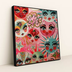 This artwork shows whimsical painted faces with large lashes and bright lips on colorful circles, surrounded by flowers and stars, in a black frame.