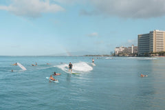 Jess Loiterton - Surfers ride waves on a sunny day at Waikiki Beach with a view of the Honolulu skyline in the background.