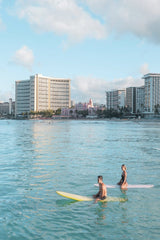 Jess Loiterton - Two surfers enjoying a sunny day on the ocean near coastal skyscrapers.
