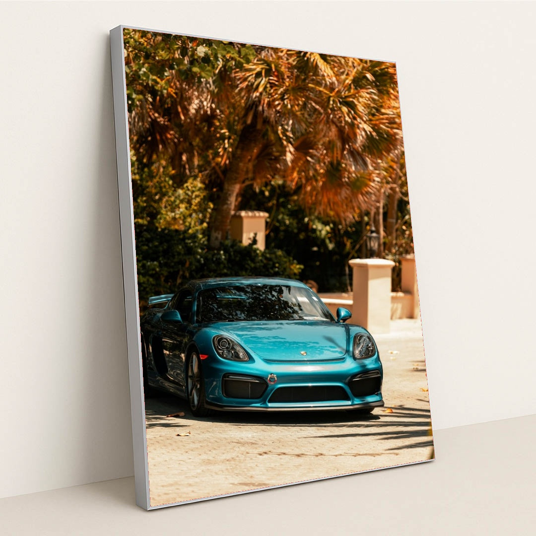 This photo shows a metallic blue Porsche sports car parked under tropical palm trees on a sunny day, in a silver frame.
