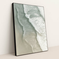 This artwork shows soft ocean waves washing onto smooth sand from above, in a black frame.