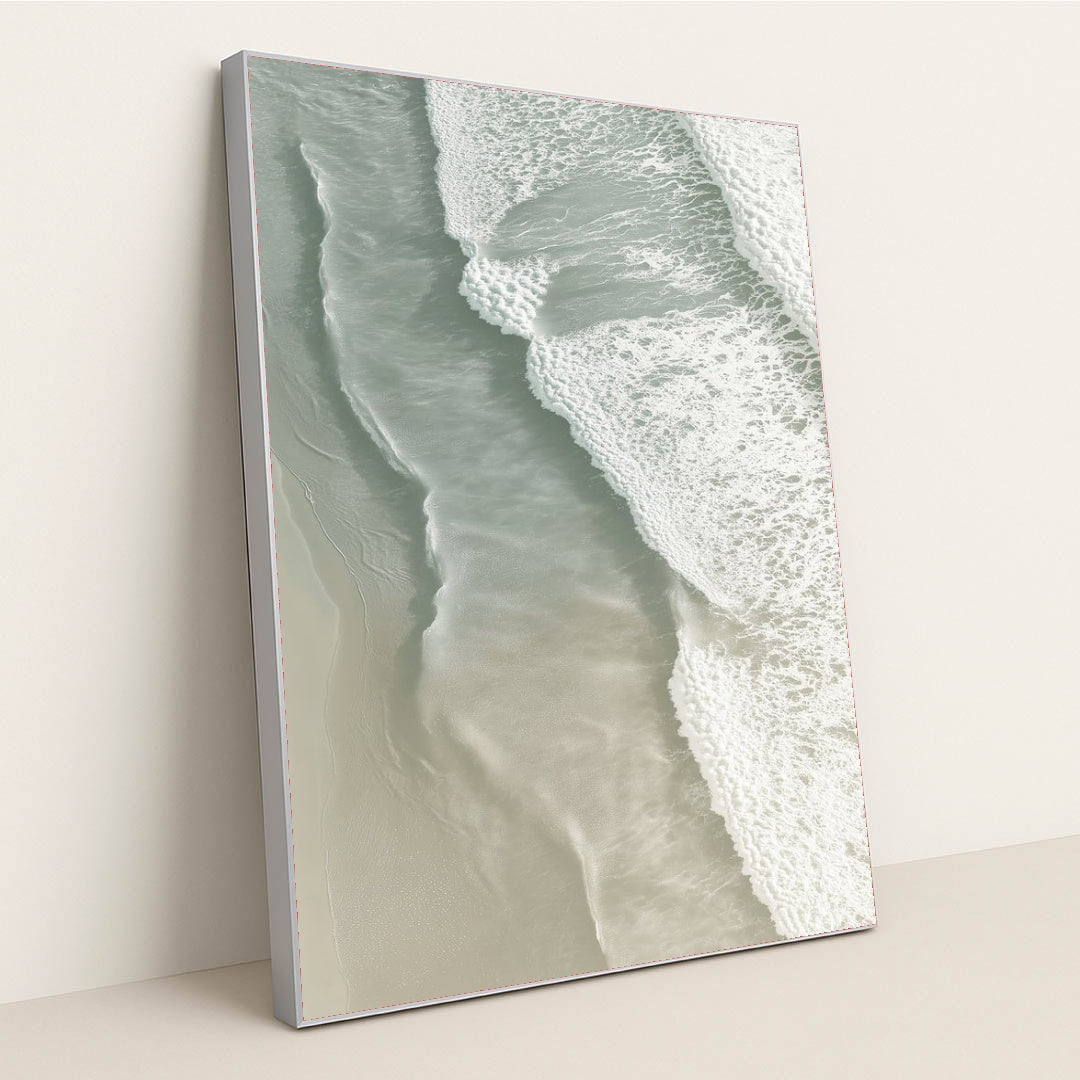 This artwork shows soft ocean waves washing onto smooth sand from above, in a silver frame.