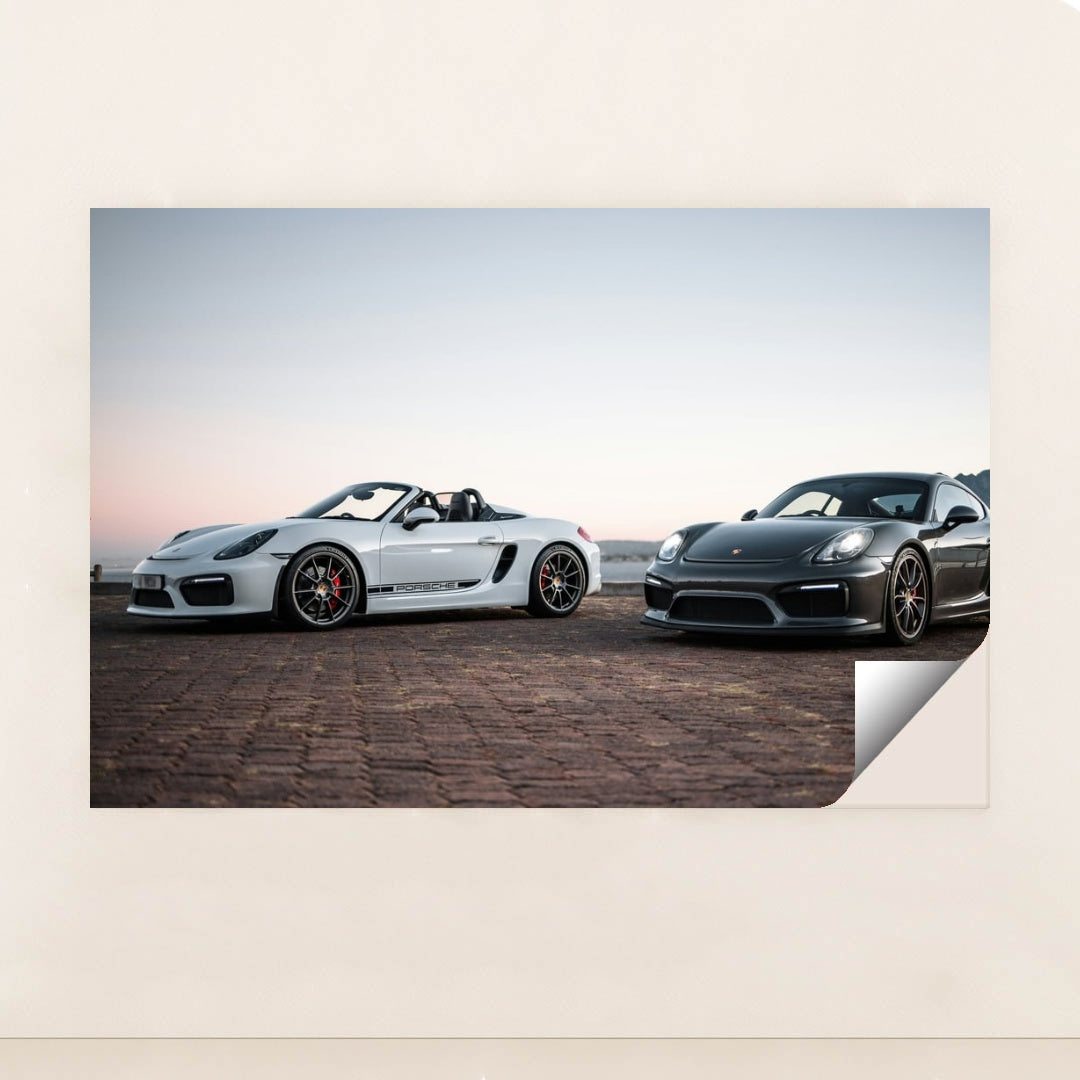 This photo shows a white convertible Porsche and a black coupe Porsche parked together at sunrise, unframed.