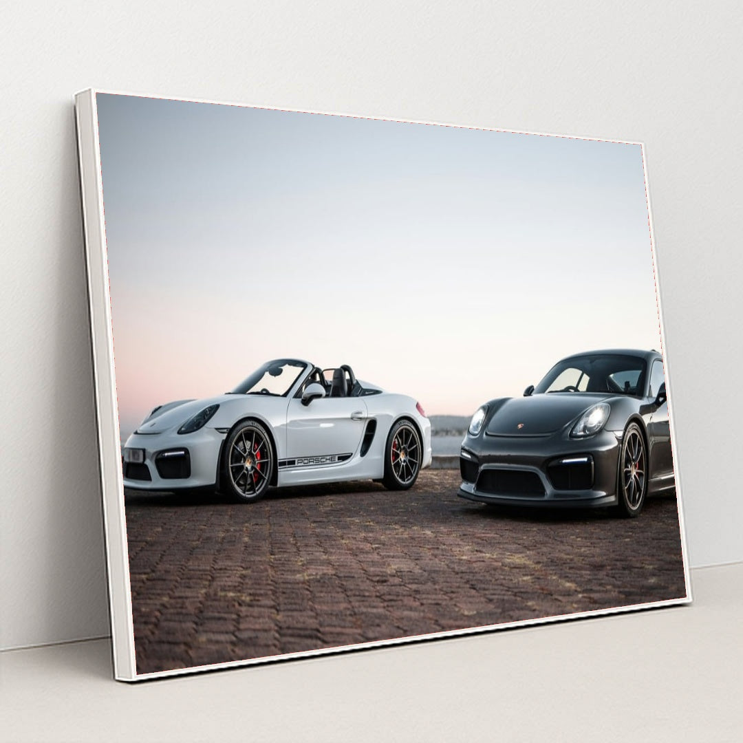 This photo shows a white convertible Porsche and a black coupe Porsche parked together at sunrise, in a white frame.